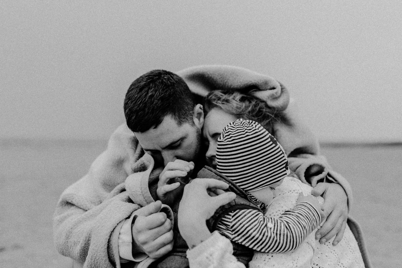 Familienfotografie Kiel — Familie umarmt sich am Strand, schwarz-weiss Geburtsreportage von Kicherbande